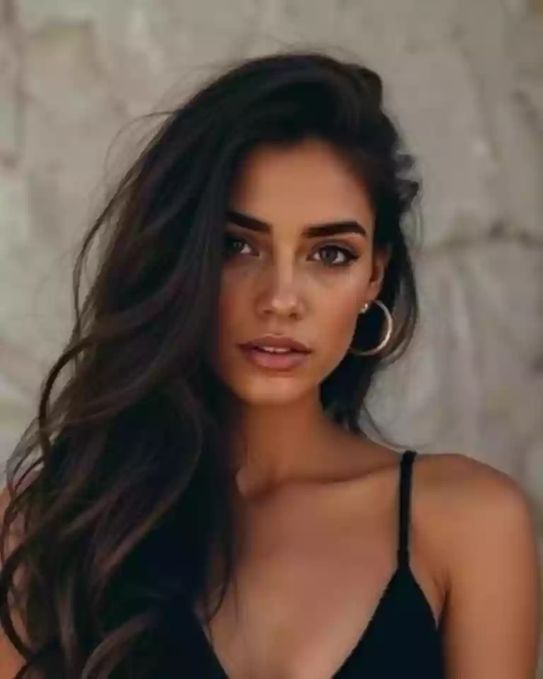 Portrait of a woman with long dark hair, wearing a black spaghetti-strap top and hoop earrings, looking at the camera against a neutral stone background.